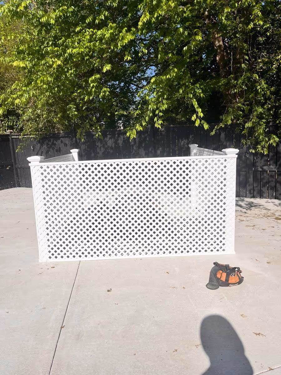 White lattice fence section placed on a concrete surface with green leafy trees and a black wooden fence in the background and a small orange and black bag on the ground.
