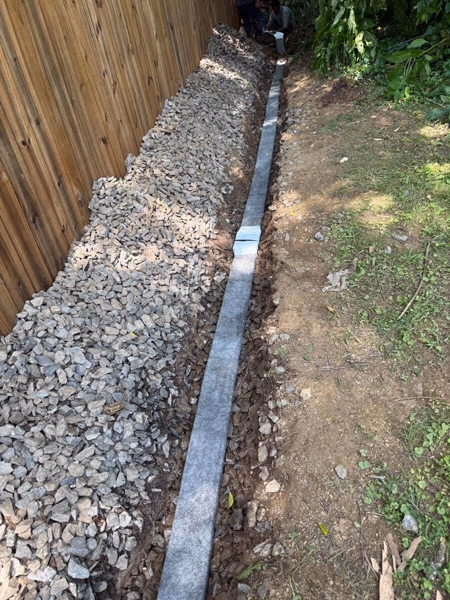 Narrow trench beside wooden fence with a layer of gravel on one side and soil on the other, and a strip of fabric or liner running through the trench with people working at the far end.