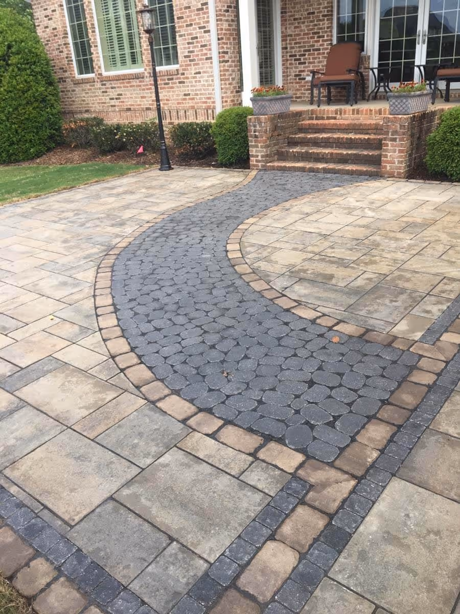Curved stone pathway bordered by square tiles leading to brick steps and a porch with chairs and potted plants.