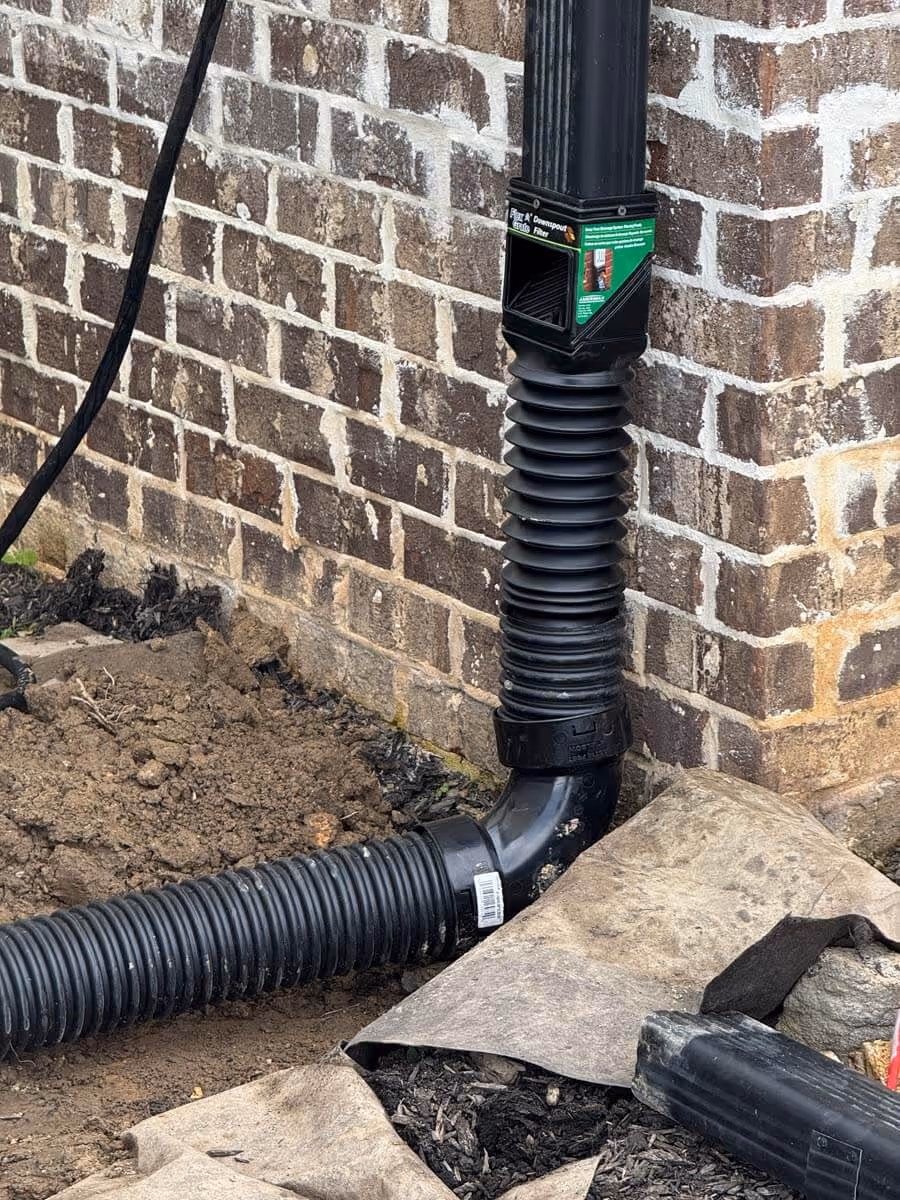 Black corrugated downspout extension attached to a brick house with soil and landscaping fabric nearby.