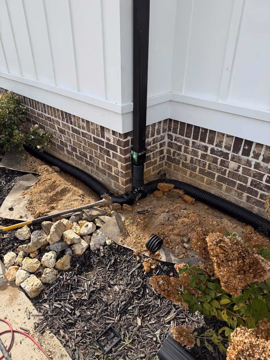 Corner of a brick house with white siding showing black corrugated drain pipes connected to a downspout and soil with rocks and garden mulch nearby.