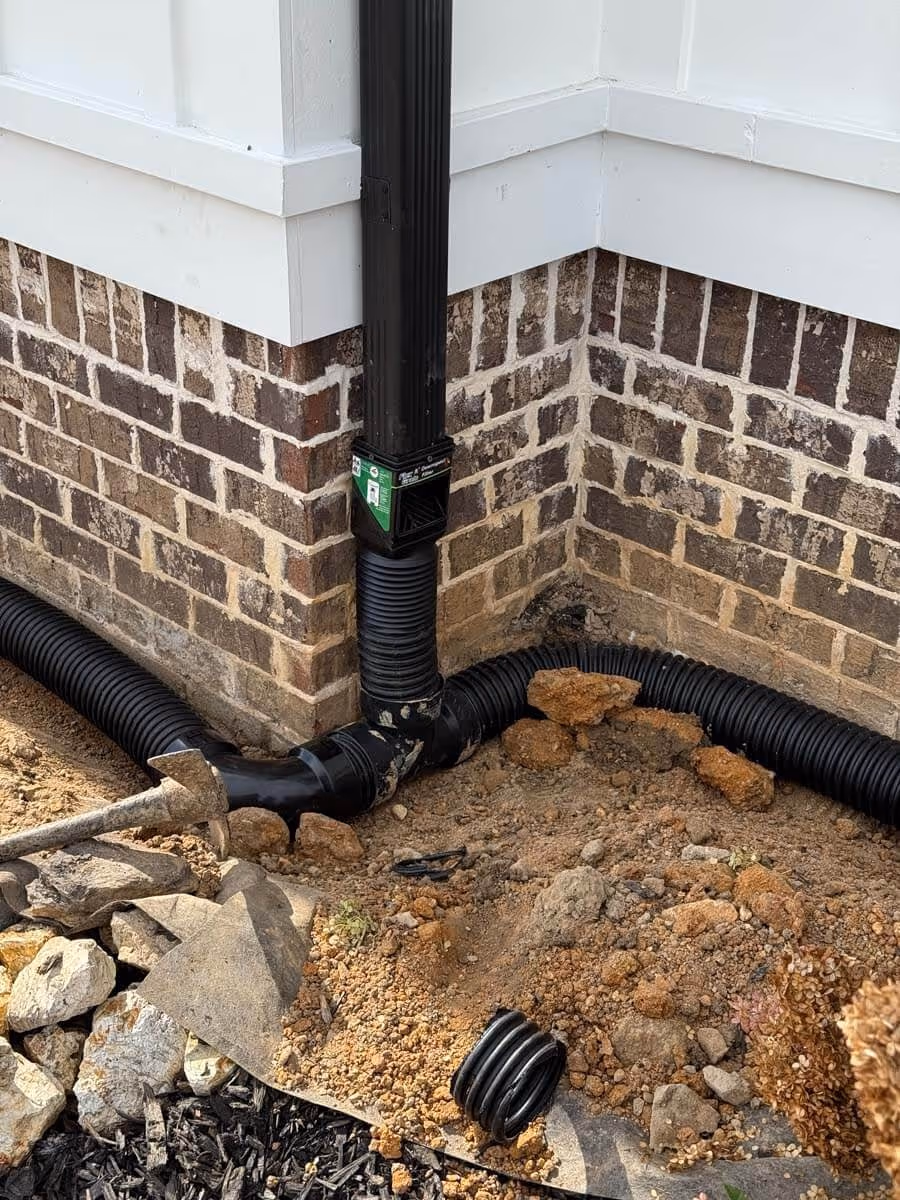 Black downspout connected to corrugated drainage pipes on the ground beside a brick and white siding house corner with some loose soil and rocks.