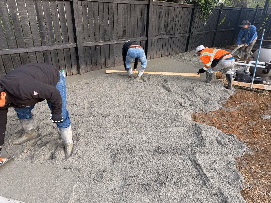 Four workers spreading and leveling freshly poured concrete in an outdoor area near a wooden fence.