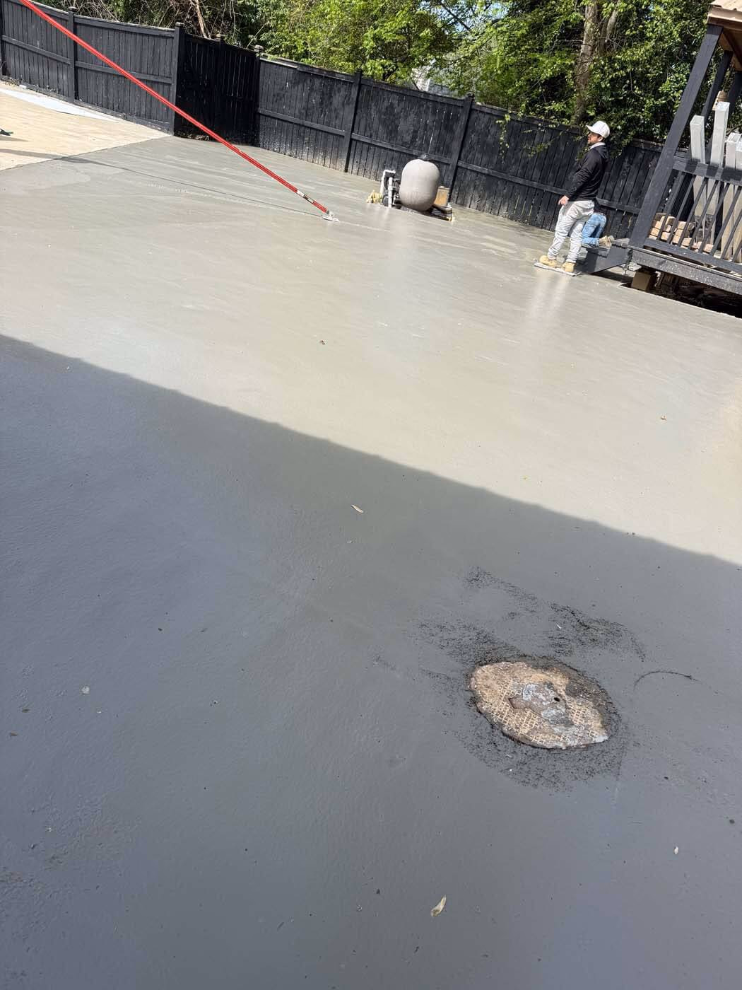 Two workers finishing a freshly poured concrete surface in an outdoor space with a black wooden fence and trees in the background.