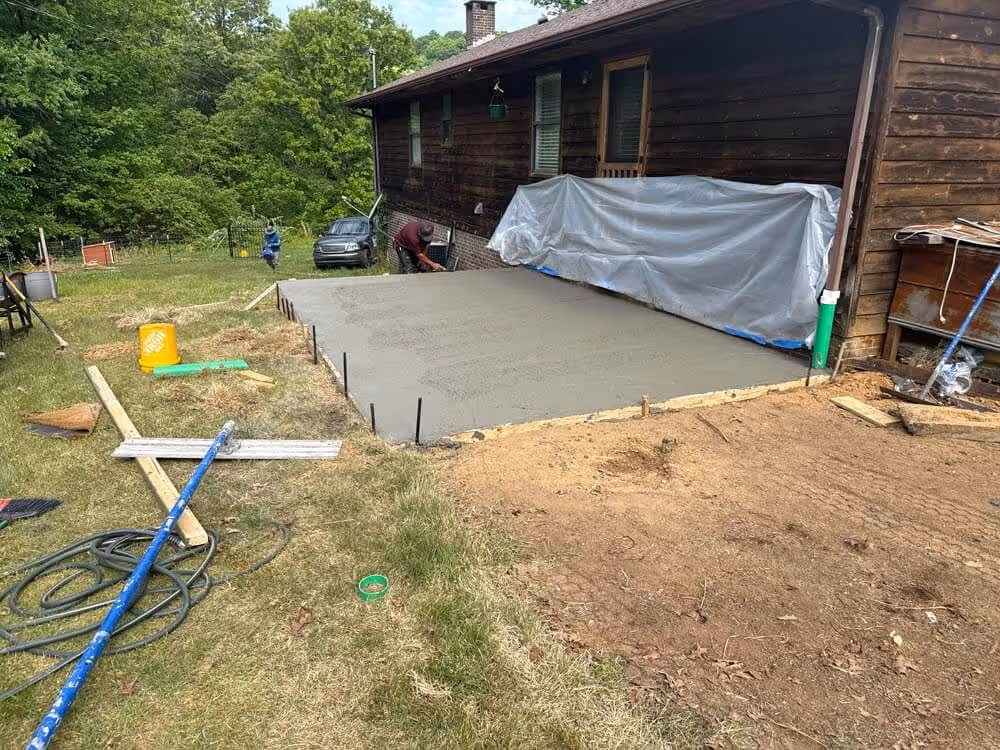 Freshly poured concrete slab next to a wooden house with construction tools and materials scattered on the surrounding grass and dirt.