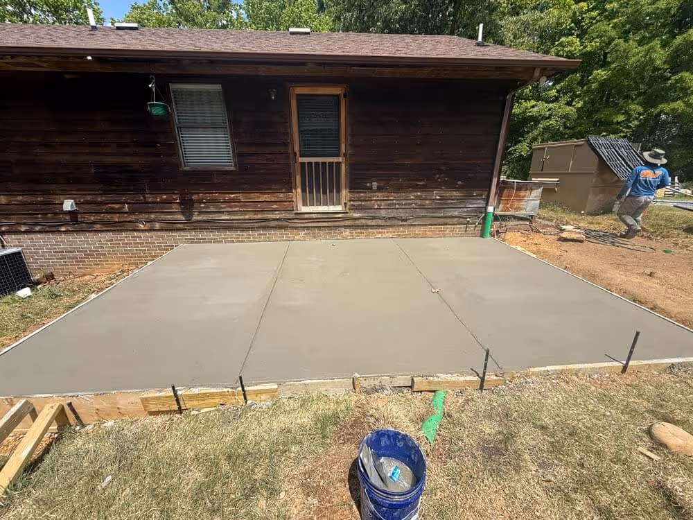 Newly poured concrete slab in front of a wooden house with a door and window, with a worker walking nearby and construction materials around.