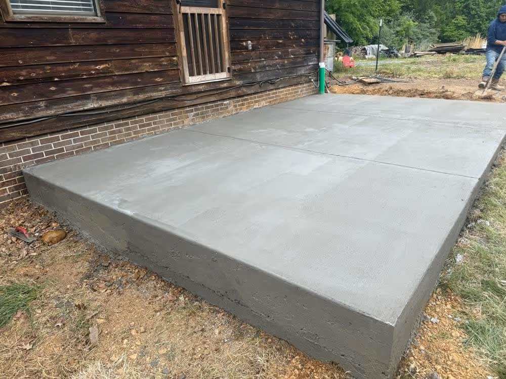 Newly poured and smoothed concrete slab next to a wooden house with a man working in the background.