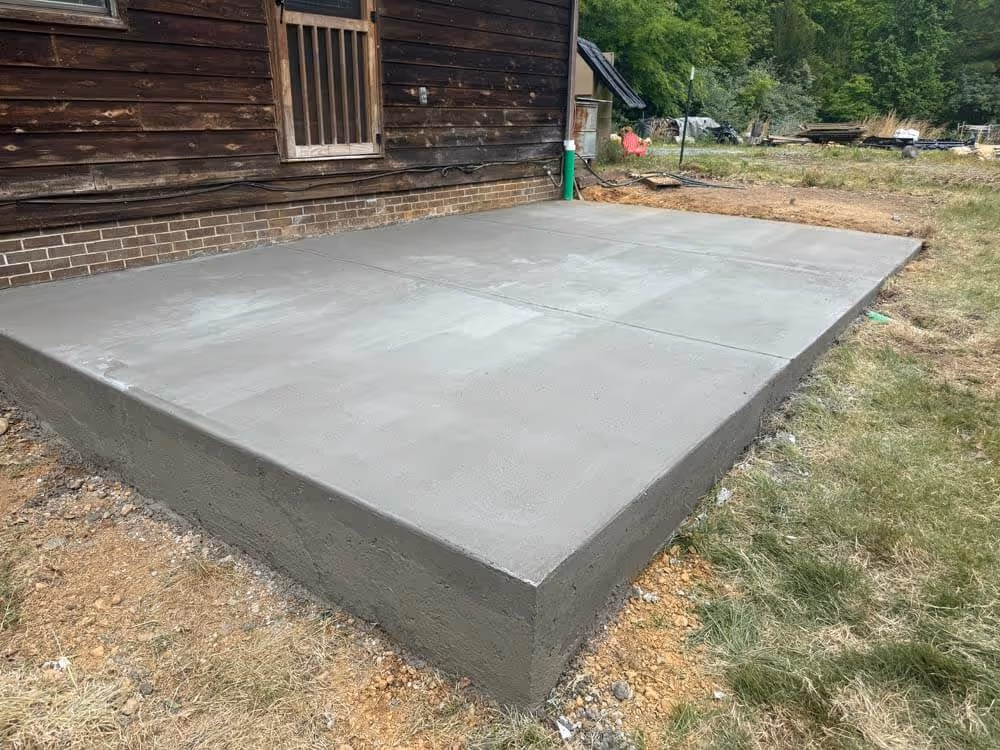 Freshly poured smooth concrete slab next to a wooden house exterior with grass and dirt surrounding it.