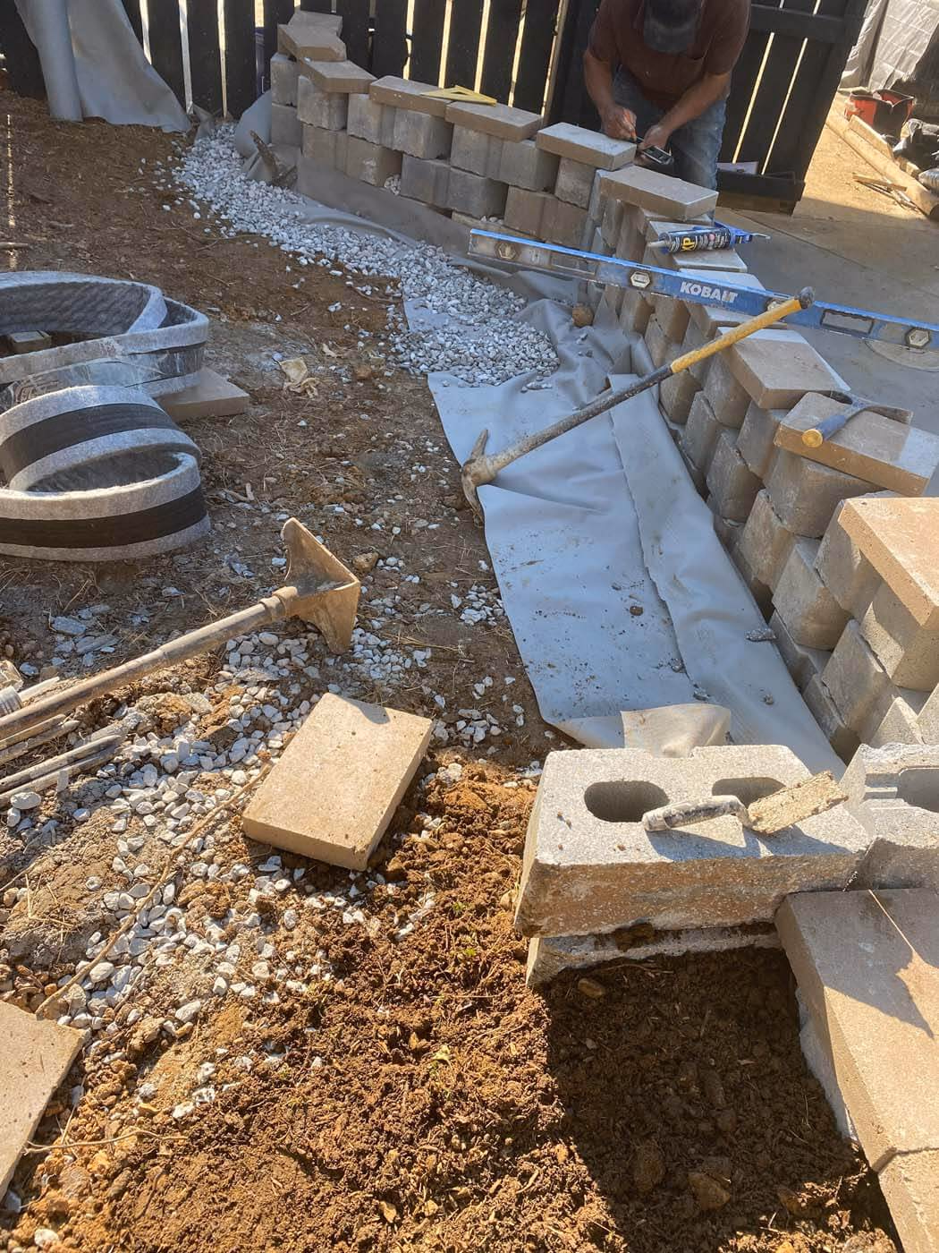 Construction site with partially built cinder block wall, tools, gravel, and fabric laid on the ground.