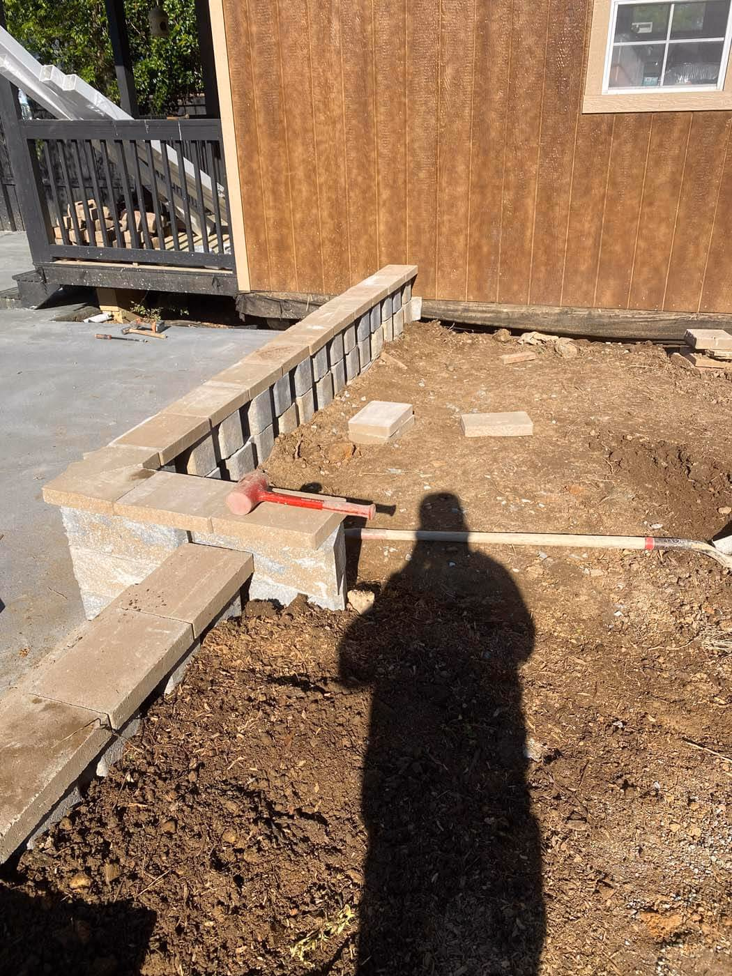 Partially constructed low retaining wall made of concrete blocks with a red mallet resting on top, next to a dirt area and a wooden building wall.