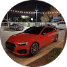 Red sports car parked on a circular brick driveway at night with streetlights and a car dealership in the background.